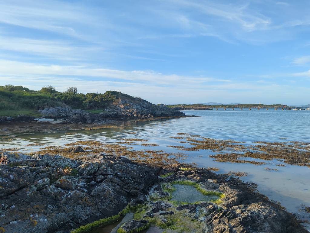 A rockpool next to the water at evening. In the distance are some boats.